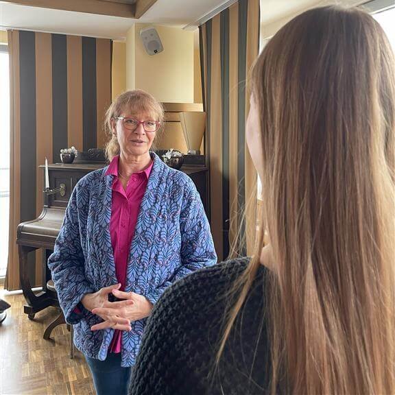 Psychologe Bettina Kappe in een adviesgesprek over het thema mental load en aandacht, staand in een ruimte met een piano en decoratieve elementen, in gesprek met een vrouw die van achteren te zien is.