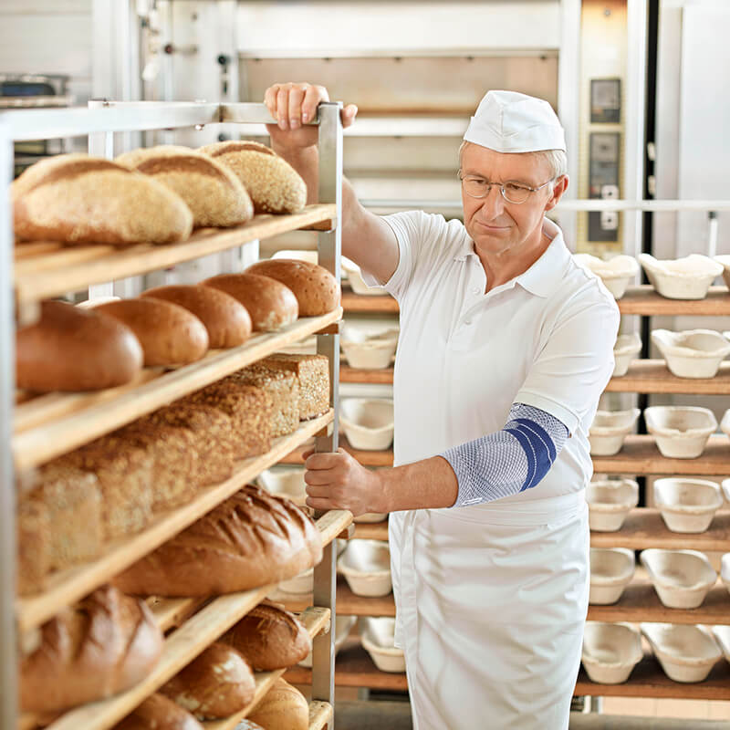 Senior bakker met Bauerfeind EpiTrain elleboogbrace bij schappen met vers brood.