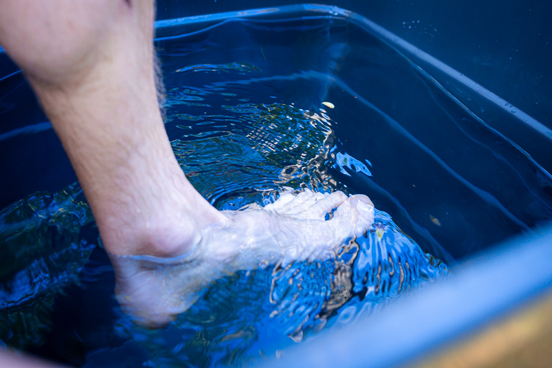 Een voet dompelt zich onder in koud water in een blauwe bak. Het heldere water vertoont lichte rimpelingen en lichtreflecties op het oppervlak.
