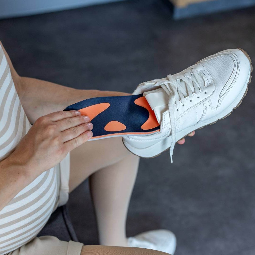 Zwangere vrouw legt een oranje-blauwe inlegzool van Bauerfeind in een witte sneaker. De inlegzool is speciaal afgestemd op de behoeften van zwangere vrouwen, om comfort en ondersteuning bij het lopen te bieden.