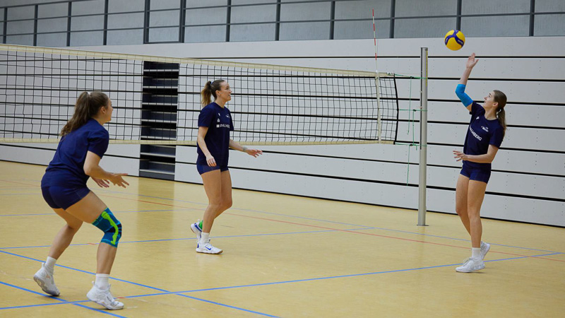 Drie volleybalsters op een indoorveld tijdens een wedstrijd. Een speelster draagt een blauw-groene kniebeugel, een andere een blauwe armsteun.