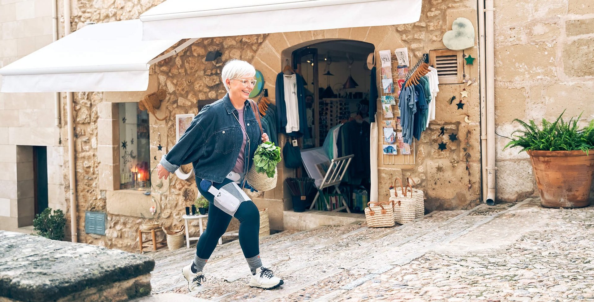 Een oudere vrouw met kort wit haar en bril loopt een geplaveid straatje omhoog. Ze draagt een spijkerjasje en een donkere legging met medische apparatuur en houdt een mandje met vers groen vast. Op de achtergrond een winkel met witte zonneschermen.