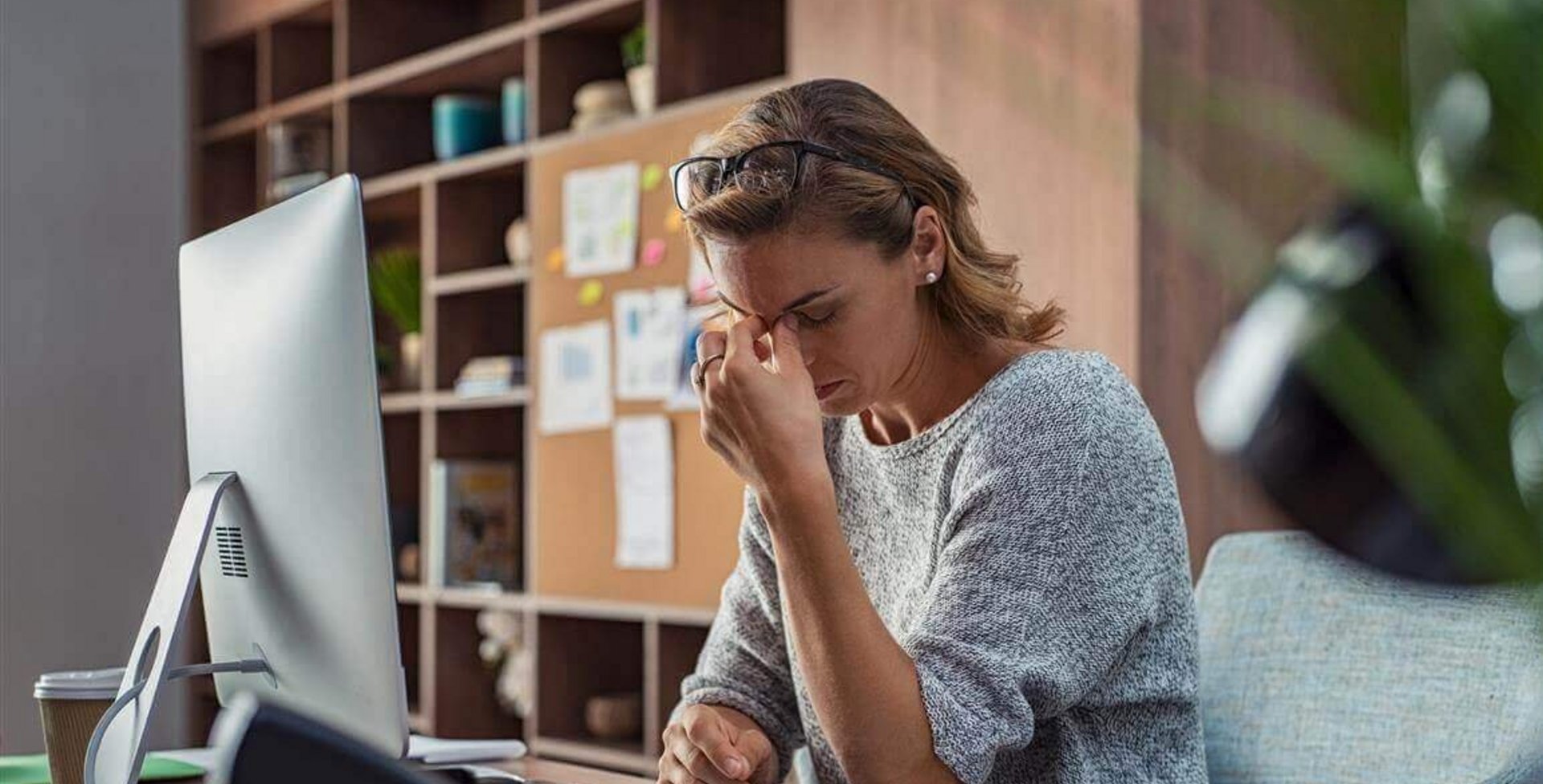 Het beeld toont een gestreste vrouw aan haar bureau op kantoor. Ze legt haar hand tegen haar voorhoofd. Te weinig beweging, bijvoorbeeld op het werk, kan ook invloed hebben op de psyche.