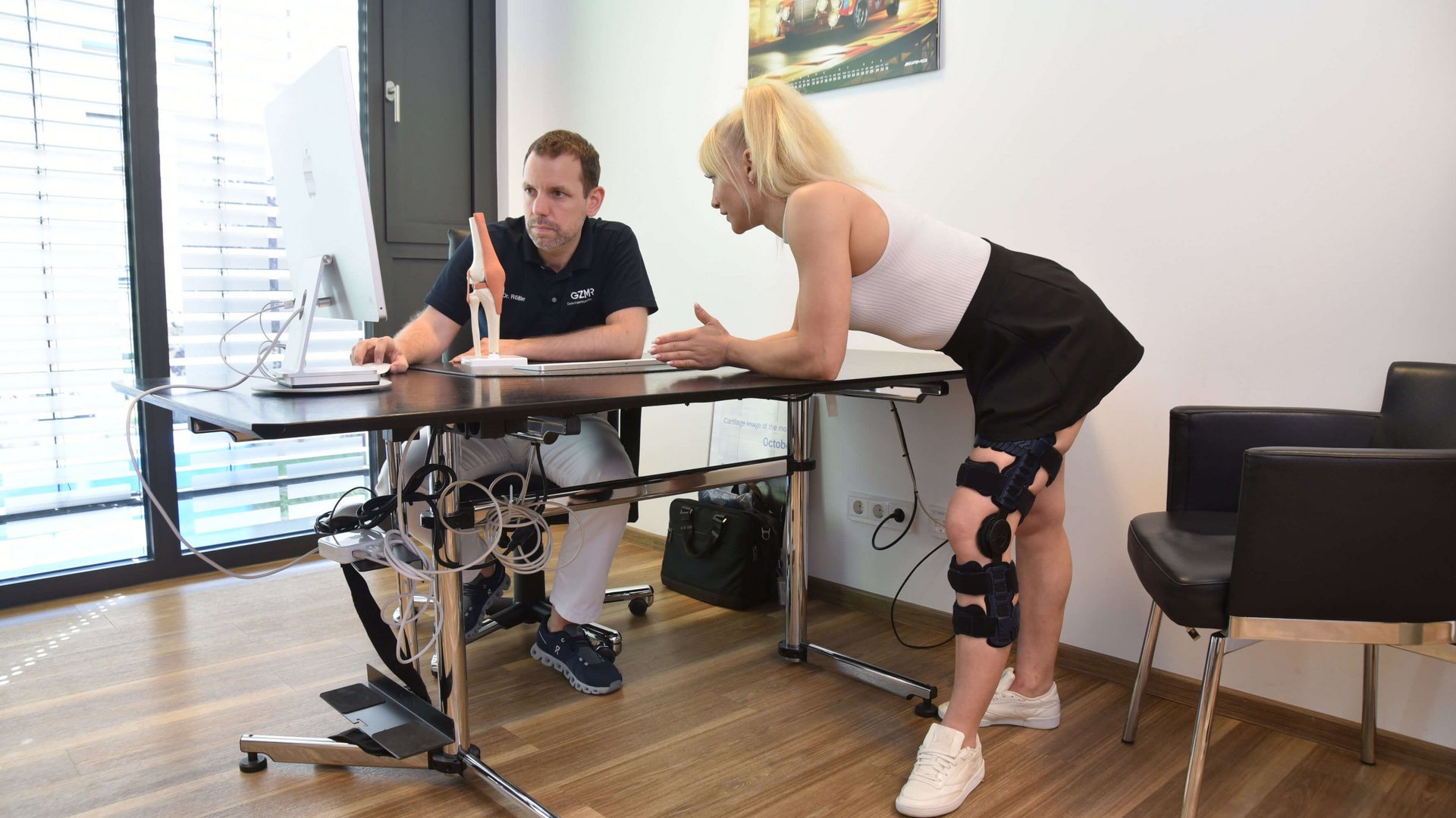 Anne-Maria Kot staat met de SecuTec Genu Flex knie-orthese bij een bureau en praat met arts Dr. Rößler, die voor een computer zit en een model van een kniegewricht laat zien.