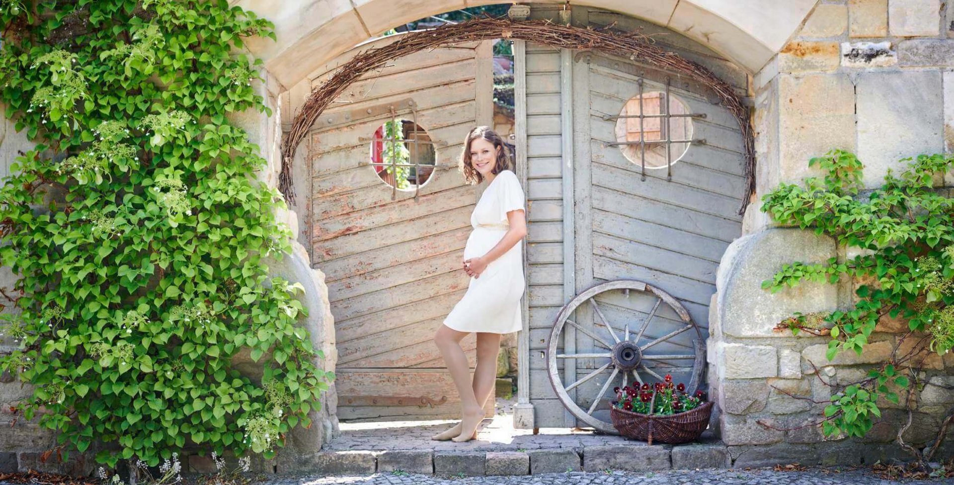 Een zwangere vrouw in een witte jurk staat voor een tuinpoort. Ze heeft haar handen op haar babybuik gelegd en lacht vriendelijk in de camera. Ze draagt VenoTrain micro compressiekousen van Bauerfeind, die de aanstaande moeder optimaal verzorgen.