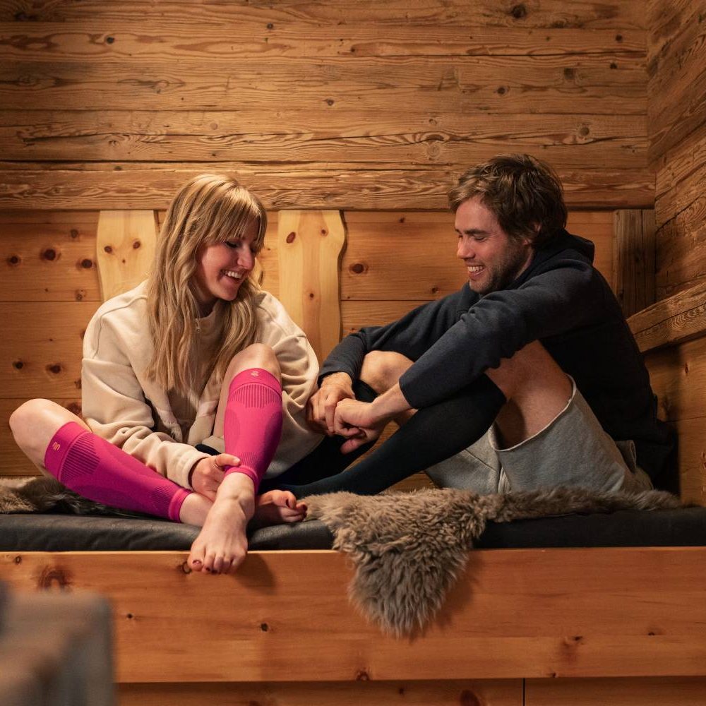 Een glimlachend stel zit gezellig in een houten hut op een bank met een bontkleed. De vrouw draagt roze compressiekousen, terwijl de man zwarte draagt. Beiden genieten van hun welverdiende rust na een actieve dag.