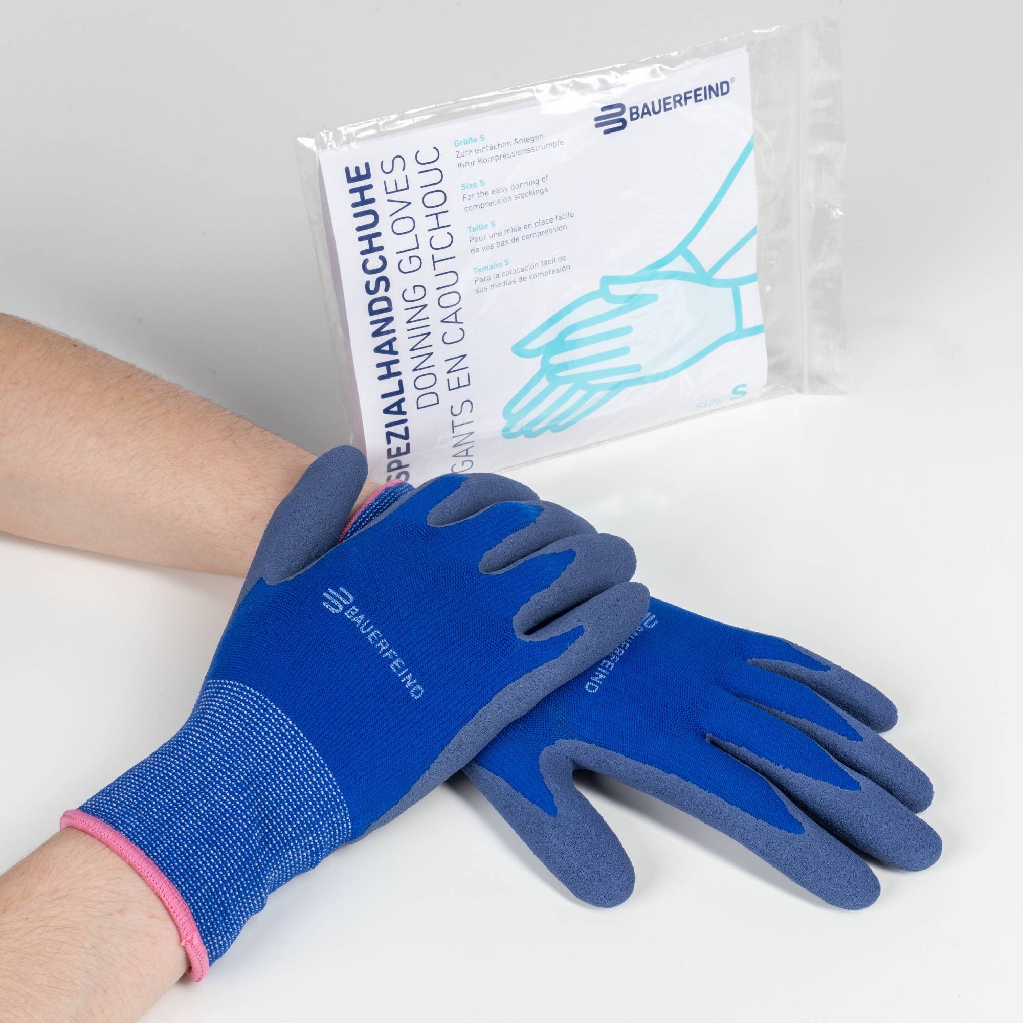 Handen met blauwe Bauerfeind aantrekhandschoenen naast verpakking met tekst ‘Donning gloves’, maat S.