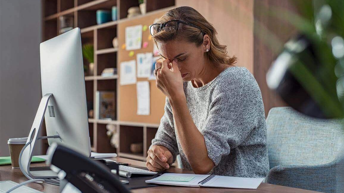 De afbeelding toont een gestreste vrouw op haar kantoorwerkplek. Ze legt haar hand tegen haar voorhoofd. Te weinig beweging, bijvoorbeeld op het werk, kan ook op de psyche inwerken.