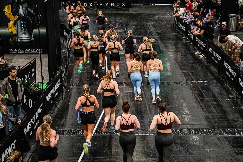 Bovenaanzicht van een HYROX-loopstrook in een indoor evenementhal, waar meerdere atleten in sportkleding over het parcours bewegen