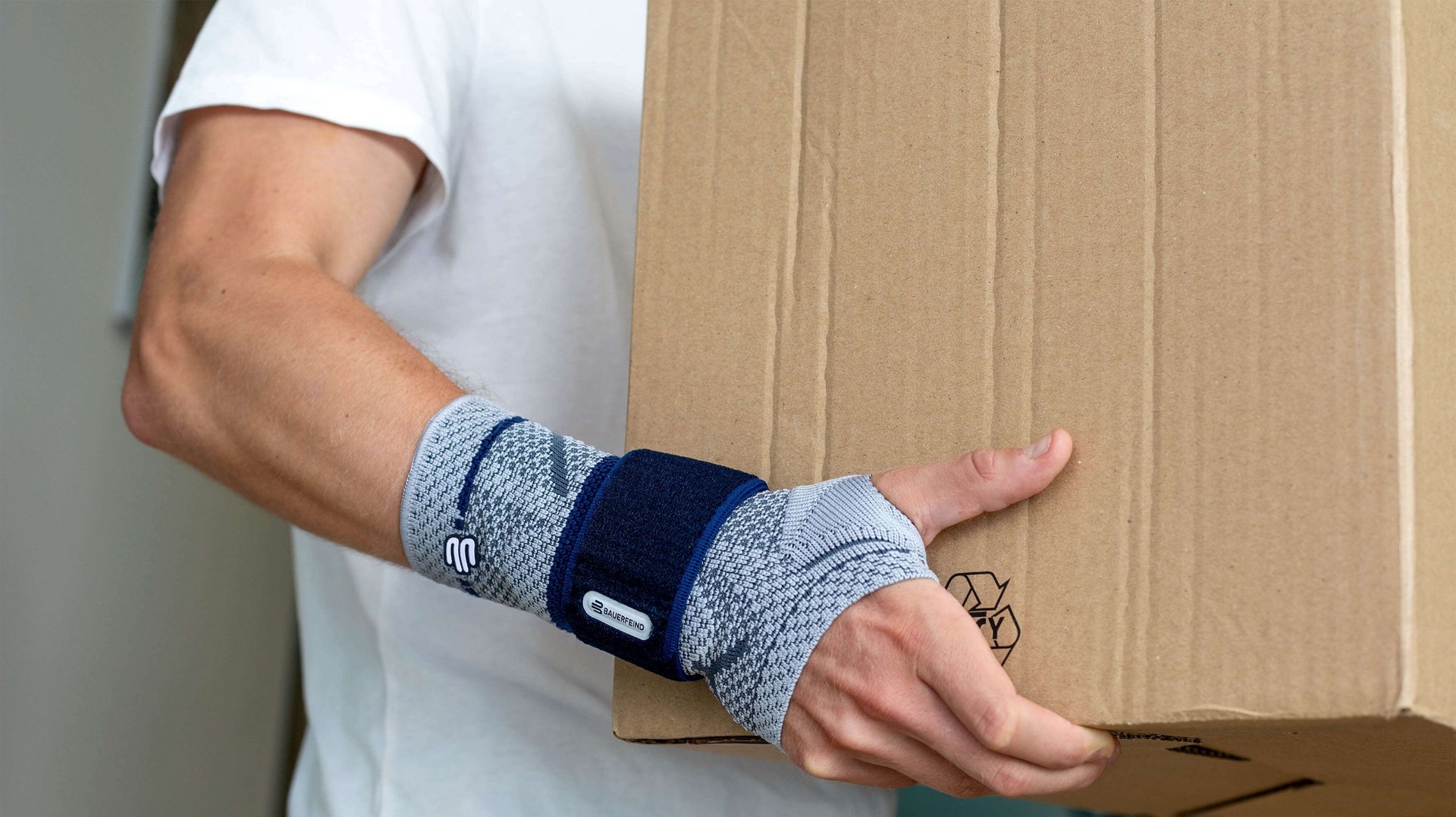 Beelduitsnede van een man die een grote doos draagt. Aan zijn rechterhand draagt hij een ManuTrain bandage van Bauerfeind. Die biedt onder andere steun bij een peesschede-ontsteking.