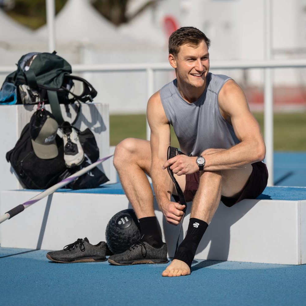 Een atleet zit op een podium bij een atletiekbaan en trekt een Bauerfeind Sports Ankle Support brace over zijn enkel. Zijn sportschoenen en een trainingsbal liggen naast hem.