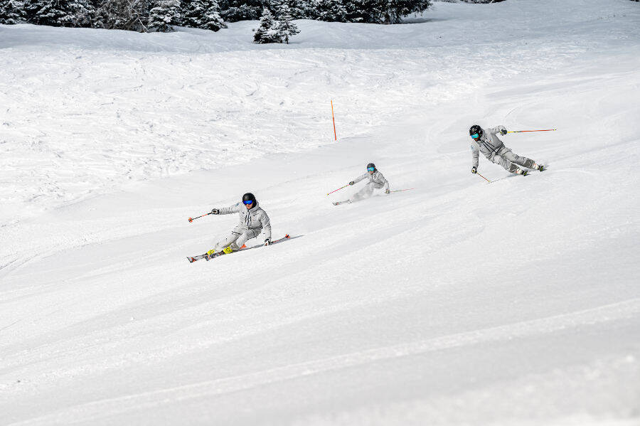 Drie skiërs gekleed in grijs grijs aan de zijkant van een gezamenlijke afdaling en in hellende rijposities.