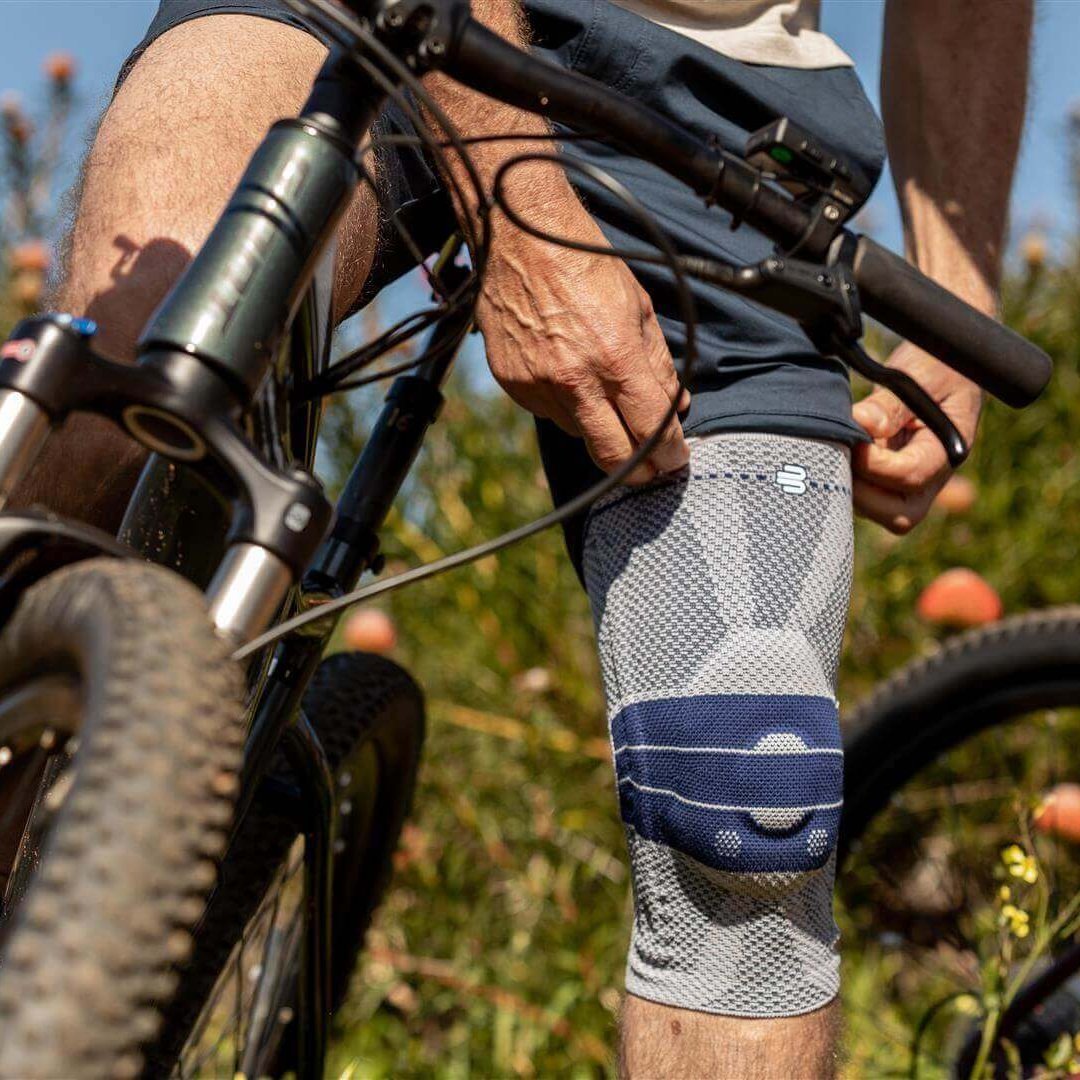 De uitsnede toont een fietser die stopt om zijn GenuTrain kniebandage aan te doen. Deze bandage biedt ondersteuning tegen pijn tijdens het fietsen.
