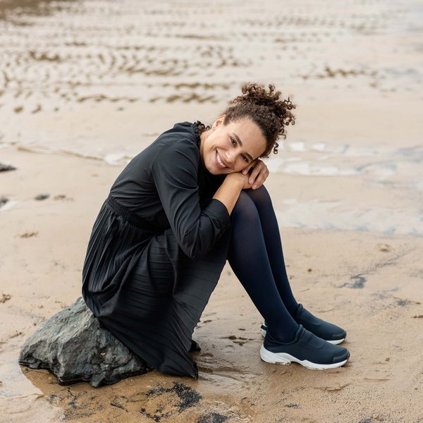 De afbeelding toont een vrouw die op een kleine rots op het strand zit. Ze draagt VenoTrain soft compressiekousen van Bauerfeind.