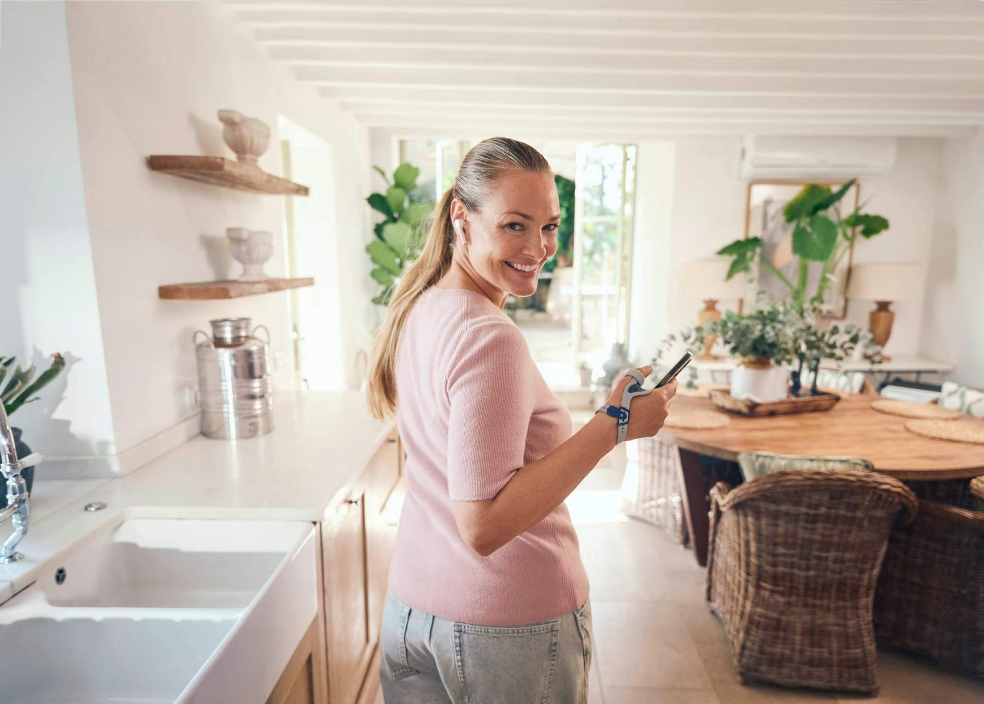 Een lachende vrouw met lang blond haar staat in een lichte, modern ingerichte keuken en houdt een smartphone in haar hand. Ze draagt een RhizoLoc OA duimorthese aan de rechterpols.