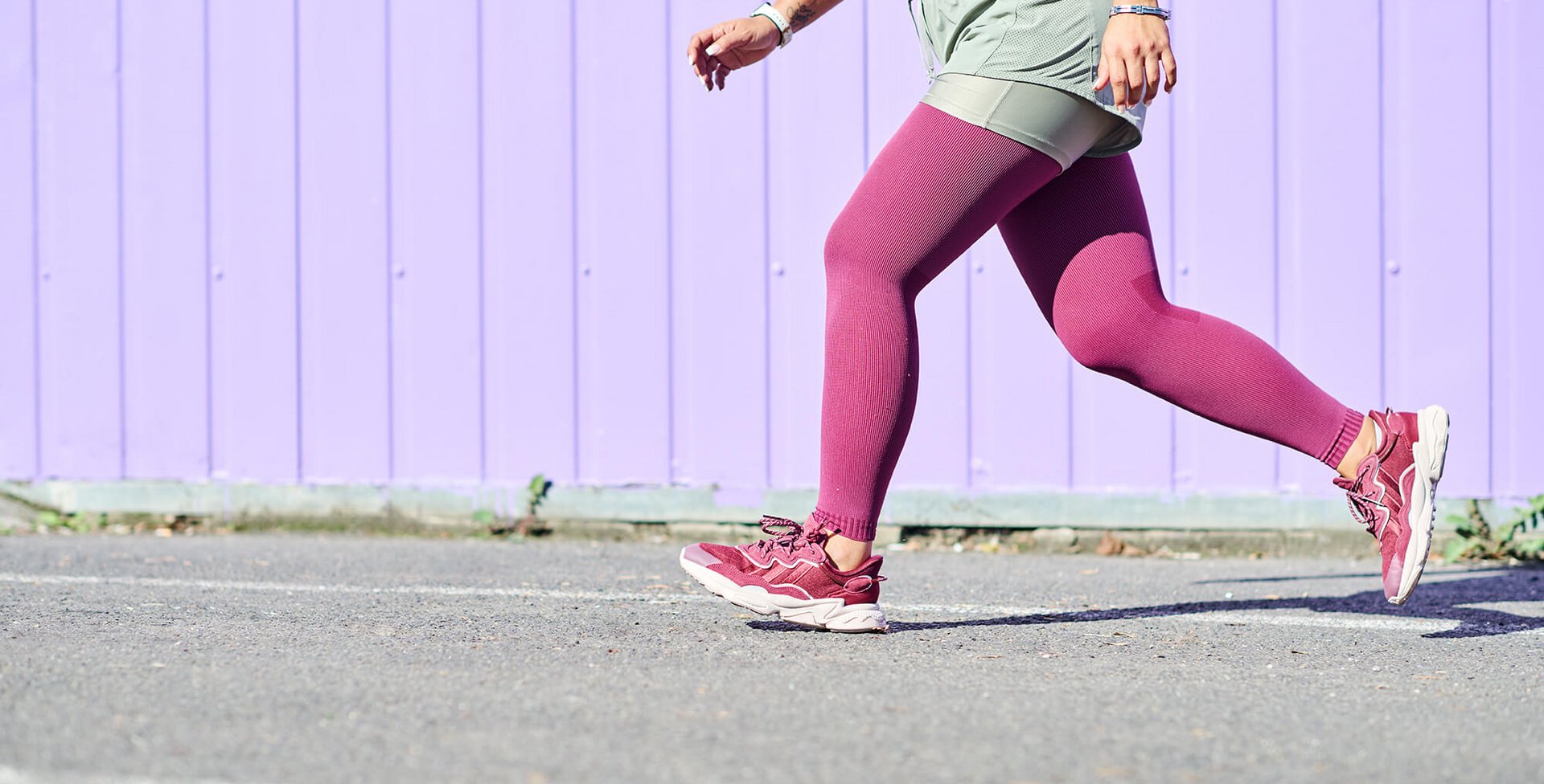 Vrouw in sportieve kleding met bordeauxrode panty's en bijpassende sneakers loopt op asfalt voor een paarse houten wand. Alleen het onderlichaam en een deel van het bovenlichaam zijn zichtbaar.
