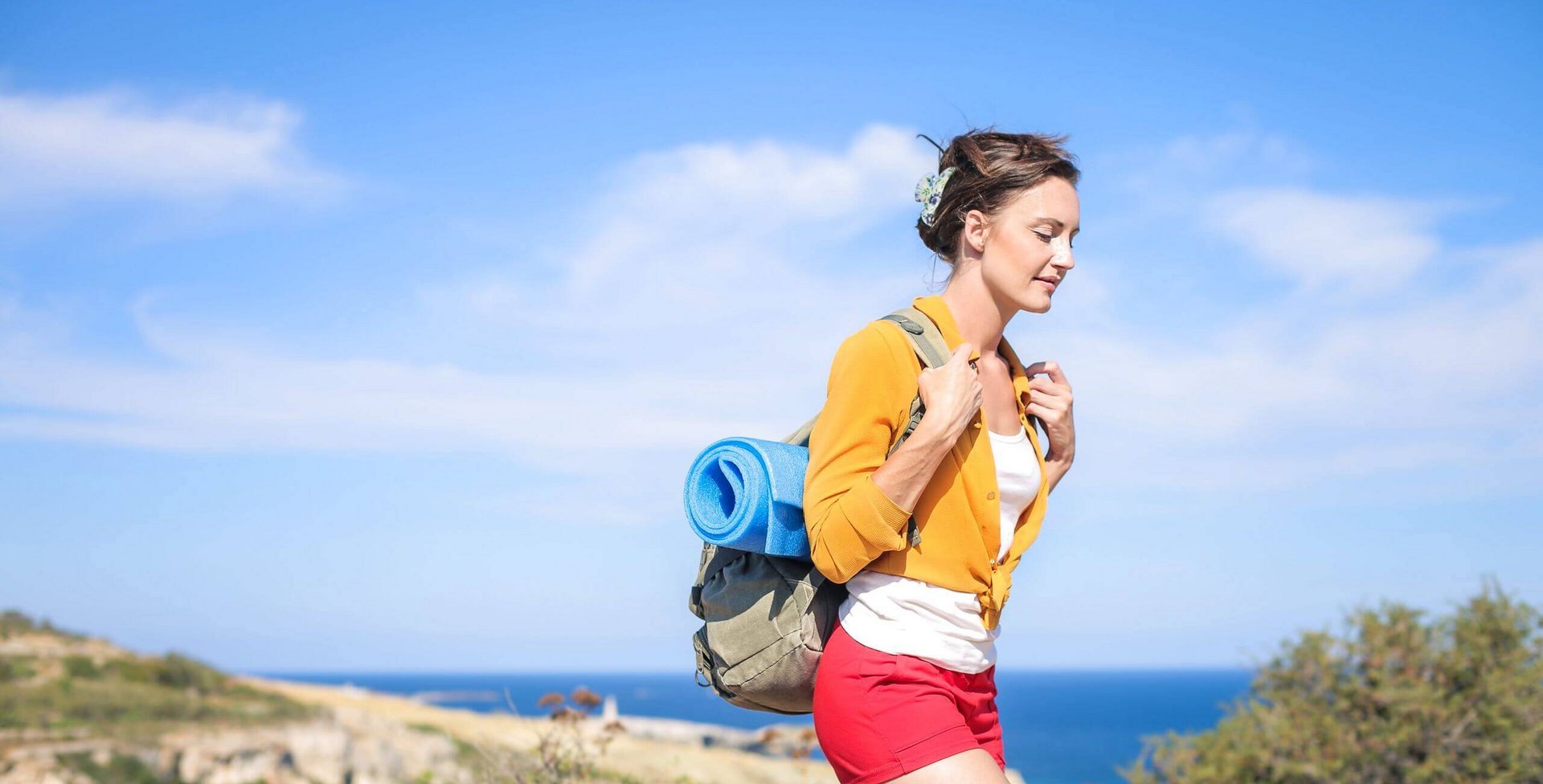 De afbeelding toont een jonge vrouw met een wandelrugzak in de zon aan zee. In de warme zomermaanden valt het ons meestal gemakkelijker om actief te worden en bewegingsgebrek tegen te gaan.