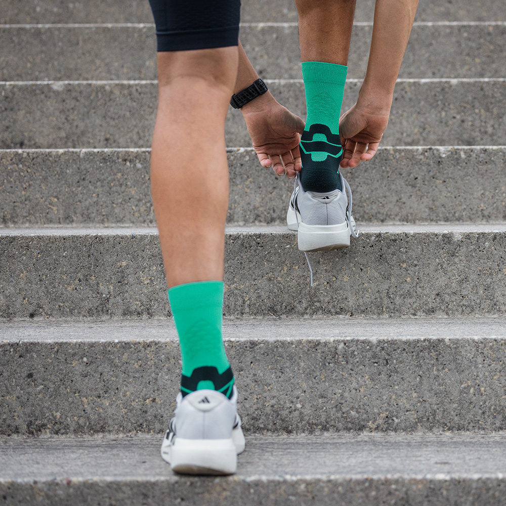 Achteraanzicht van een hardloper die een trap oploopt met groene Bauerfeind-hardloopsokken in sportschoenen. De focus ligt op de hiel en het anatomische ontwerp van de sok.