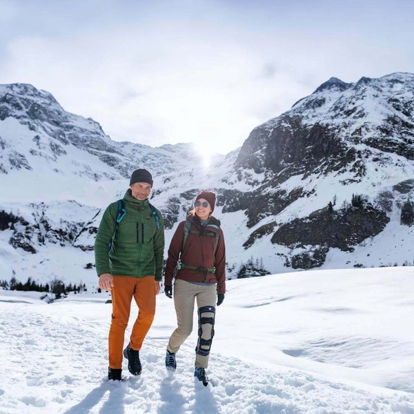 Op de foto zijn twee personen tijdens een winterwandeling in de bergen te zien. Een van deze personen draagt de knieorthese SecuTec Genu van Bauerfeind.