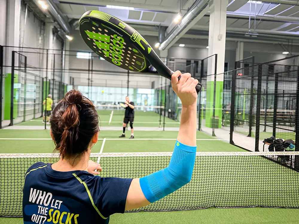 Een padelspeelster staat bij het net en maakt zich klaar om de bal te slaan. Haar arm is beschermd met een lichtblauwe compressiemouw.