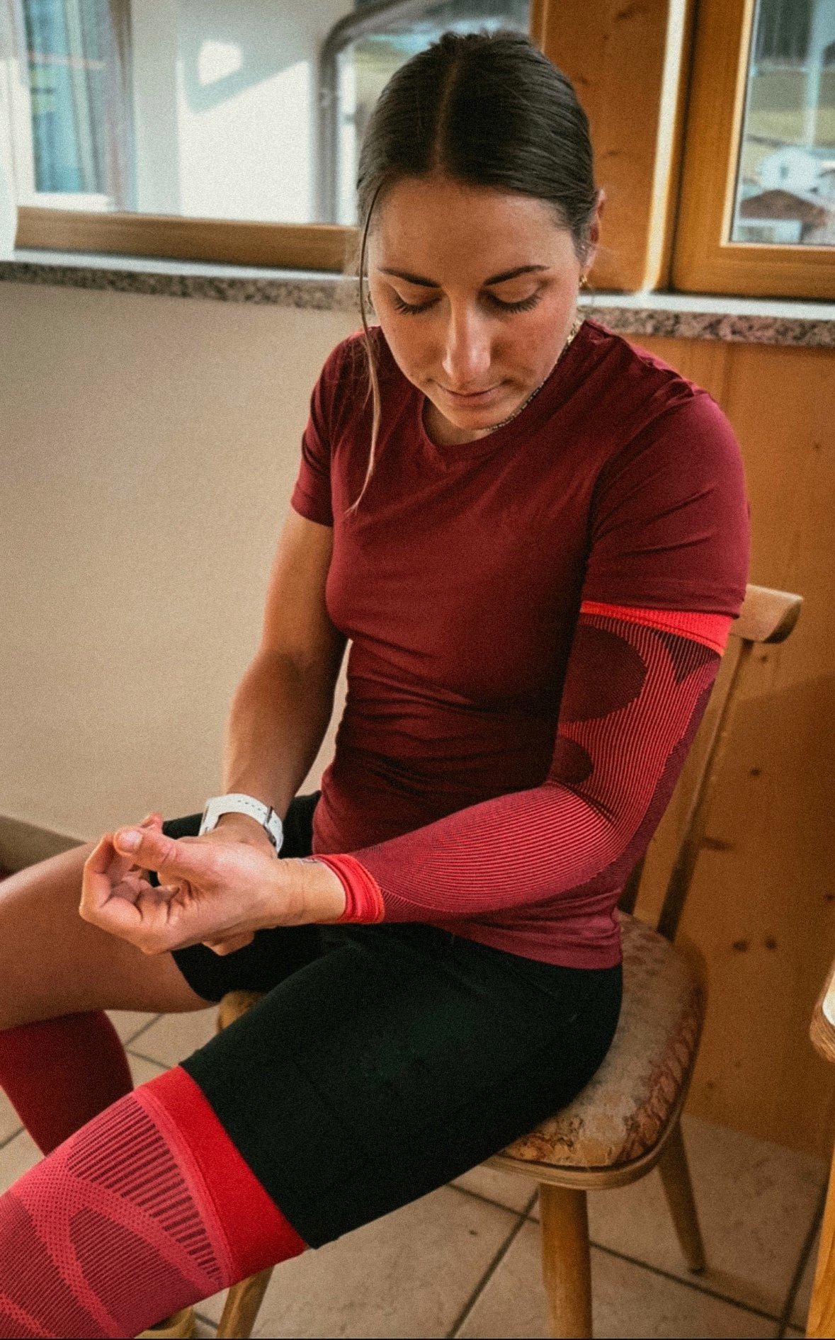 Antonia Horn zit in sportkleding op een stoel en draagt Compression Sleeves van Bauerfeind Sports om armen en benen. Ze richt haar blik naar beneden en controleert de pasvorm van de armcompressie.