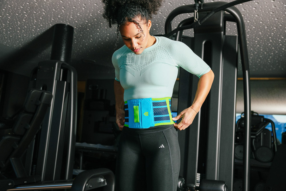Een sportieve vrouw staat in de fitnessruimte en stelt een blauwe rugbandage af rond haar taille, terwijl ze zich voorbereidt op een krachttraining.