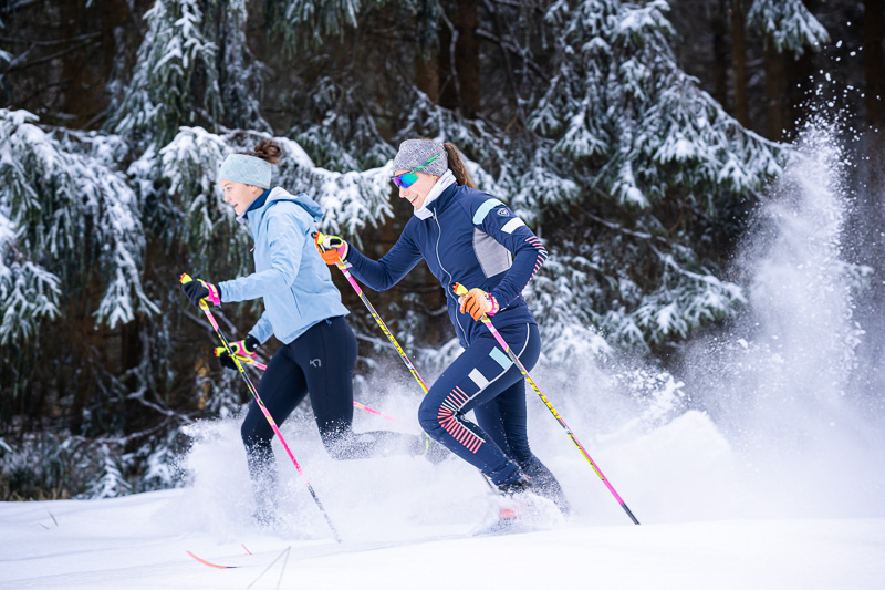 Twee vrouwen tijdens het langlaufen door met sneeuw bedekte bossen. Ze dragen kleurrijke wintersportkleding en glijden dynamisch door de sneeuw. De scène wordt gekenmerkt door dicht met sneeuw bedekte sparren, die het winterwonderland benadrukken.