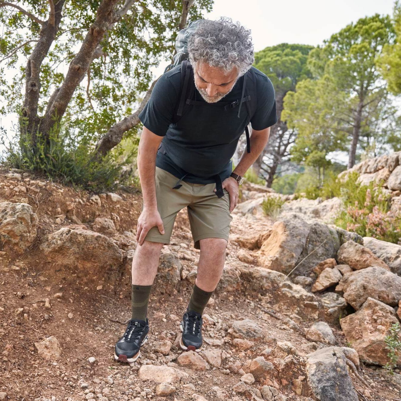 Een oudere man met grijze krullen en een rugzak houdt op een rotsachtig wandelpad stil en grijpt naar zijn pijnlijke knie. Hij draagt een zwart T-shirt, een beige korte broek en wandelschoenen.