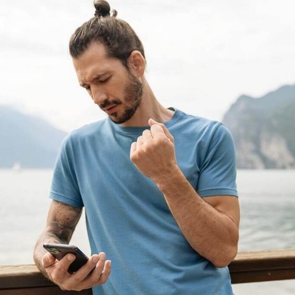 De afbeelding toont een man met een smartphone die aan de oever van een bergmeer staat. Hij houdt zijn rechterarm met pijnlijke pols omhoog.