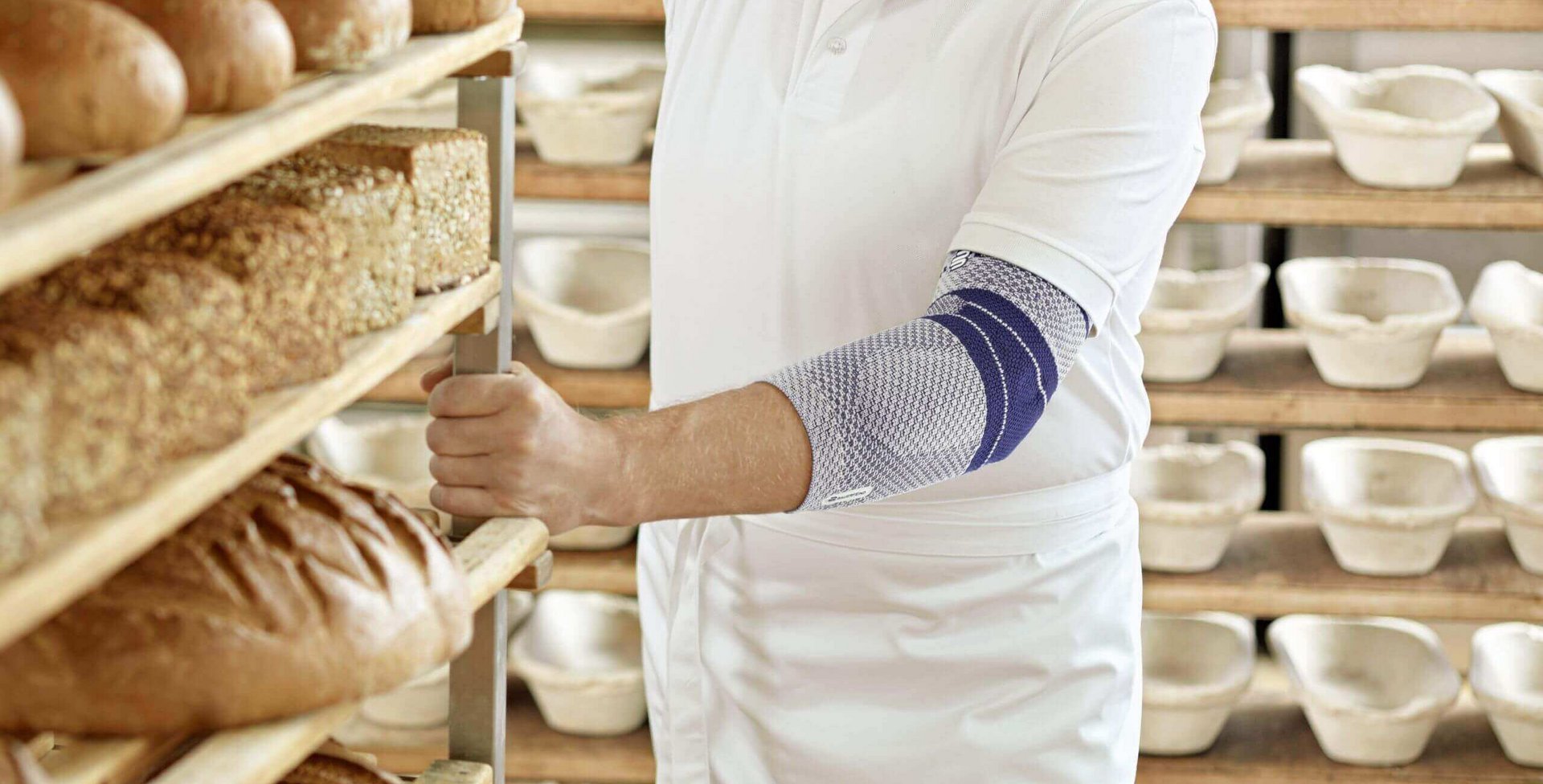 Bijgesneden beeld van een bakker in zijn bakkerij. Hij draagt een EpiTrain elleboogbandage van Bauerfeind om zijn linkerarm. De EpiTrain bandage verlicht pijn in de elleboog en verbetert de beweeglijkheid ervan.