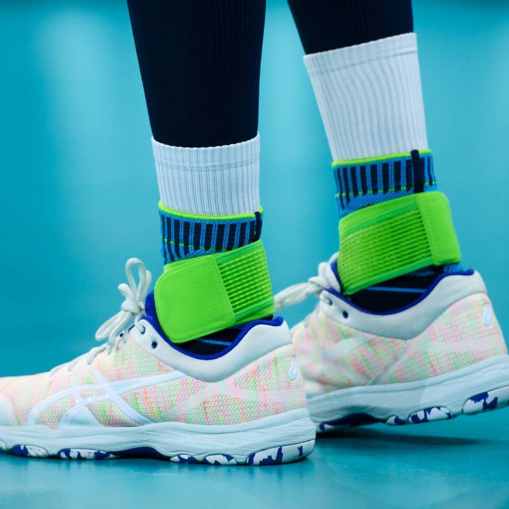 Close-up van de benen van een volleyballer op de indoorvloer. De Bauerfeind Sports Ankle Support Rivera wordt gedragen over hoge witte sokken.