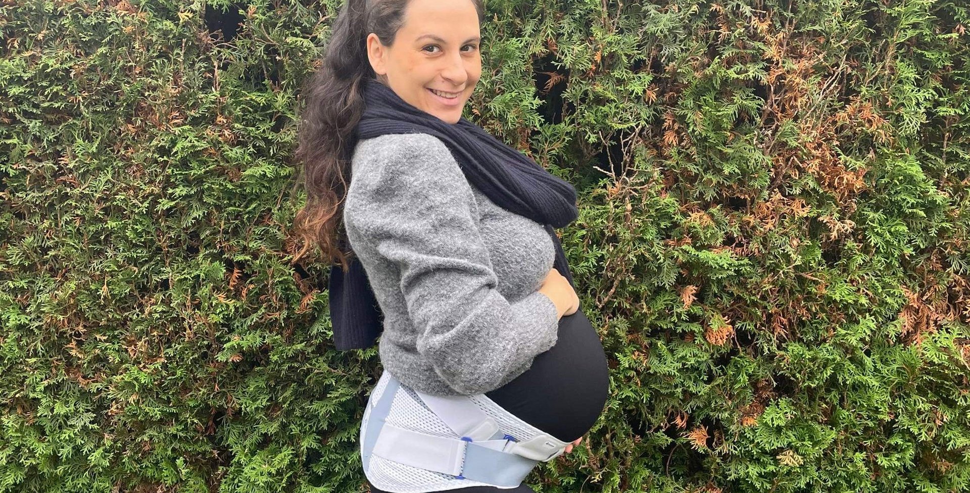 Zwangere vrouw staat in profiel voor een heg, draagt een grijze jas, een zwarte sjaal en een witte zwangerschapsorthese om haar buik.