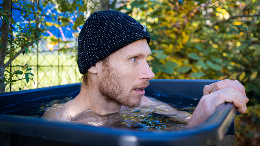 Een man zit in een zwarte waterbak, omringd door een herfsttuin. Hij draagt een zwarte gebreide muts en kijkt geconcentreerd opzij. Het water komt tot aan zijn borst en de bak staat buiten.