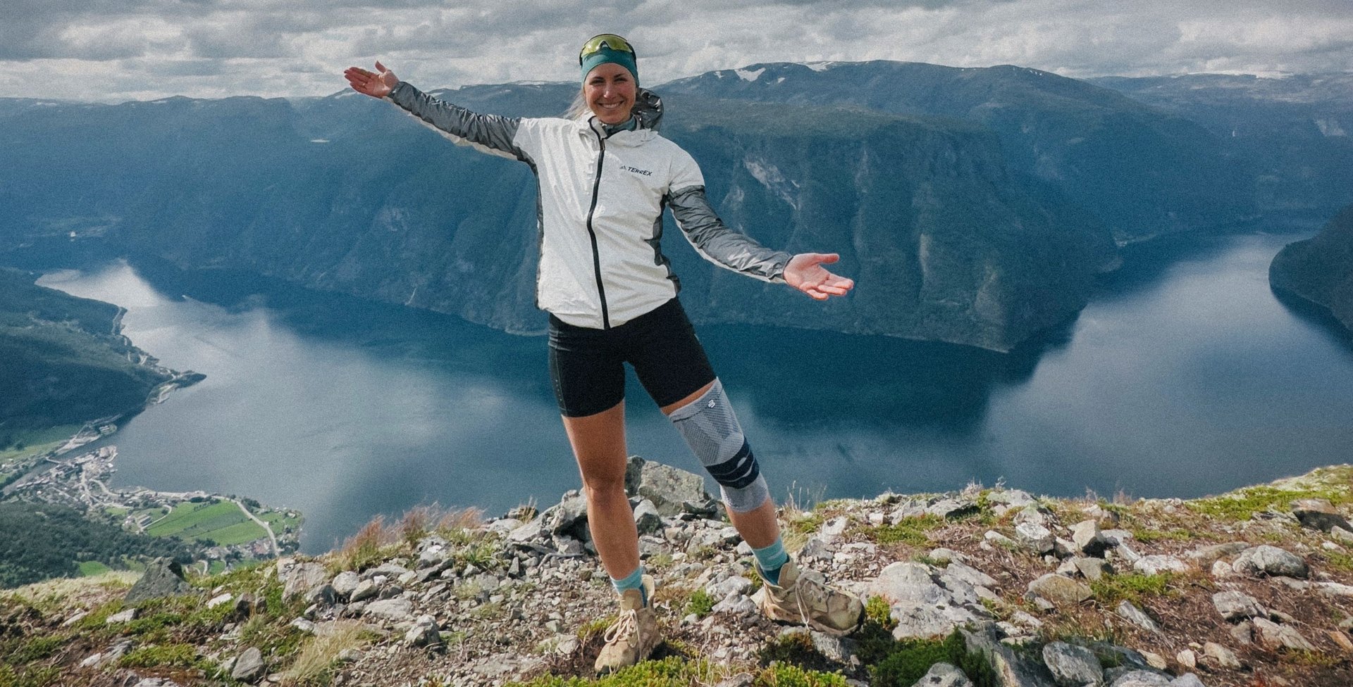 Antonia Horn staat lachend op een bergtop met gespreide armen. Ze draagt een GenuTrain-kniebandage van Bauerfeind om haar rechterknie. Op de achtergrond strekt zich een adembenemend landschap uit met fjorden en bergen onder een deels bewolkte hemel.