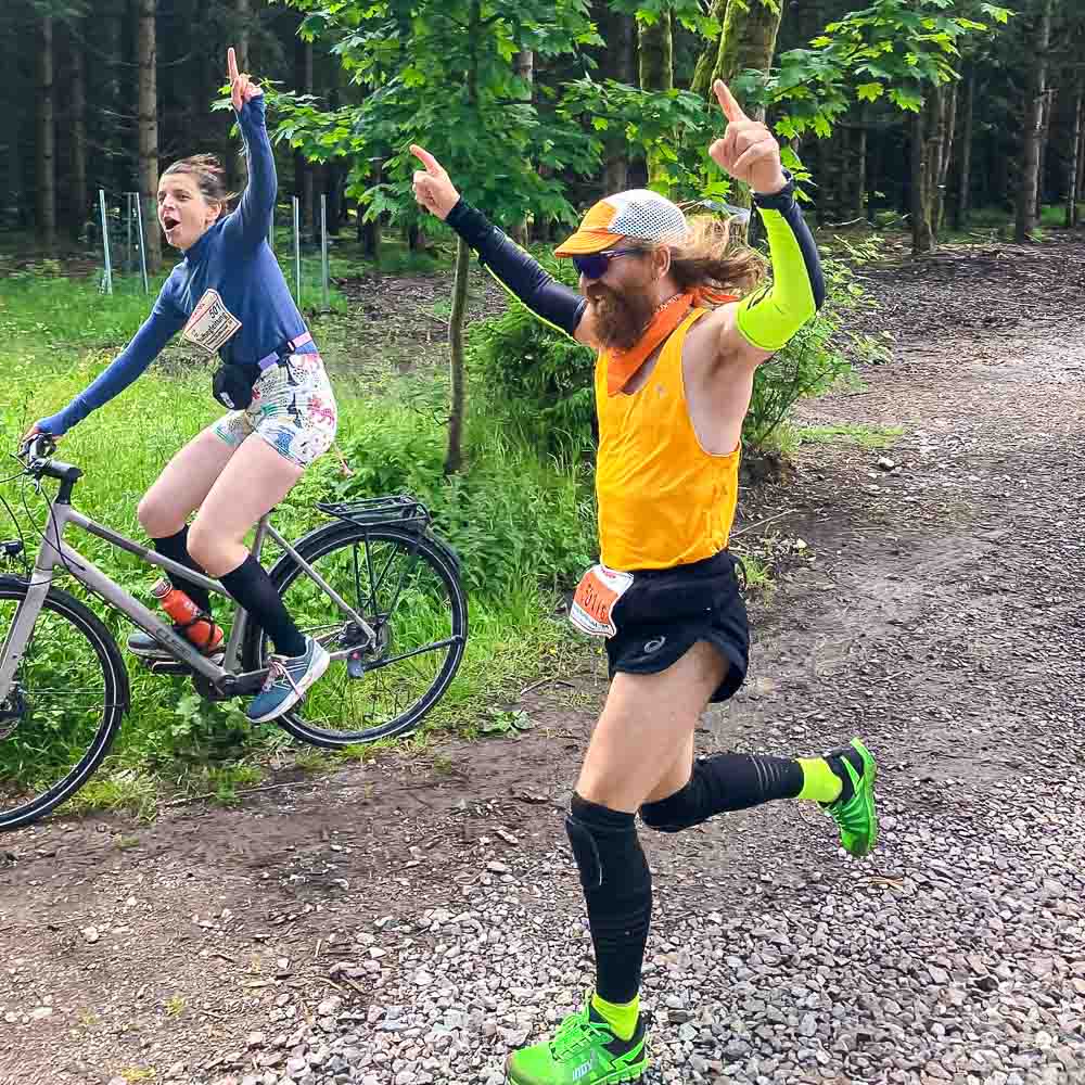 Een man rent juichend naar de finish terwijl een vrouw juichend naast hem fietst
