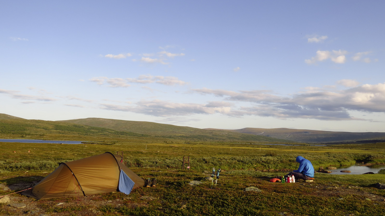 Man met een jas met een kap zit voor een tent in een landschap voor een blauwe hemel