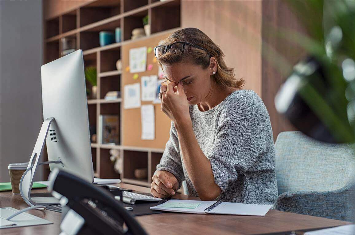 De foto toont een gestreste vrouw aan haar kantoorwerkplek. Ze legt haar hand tegen haar voorhoofd. Te weinig beweging, bijvoorbeeld op het werk, kan ook gevolgen hebben voor de psyche.