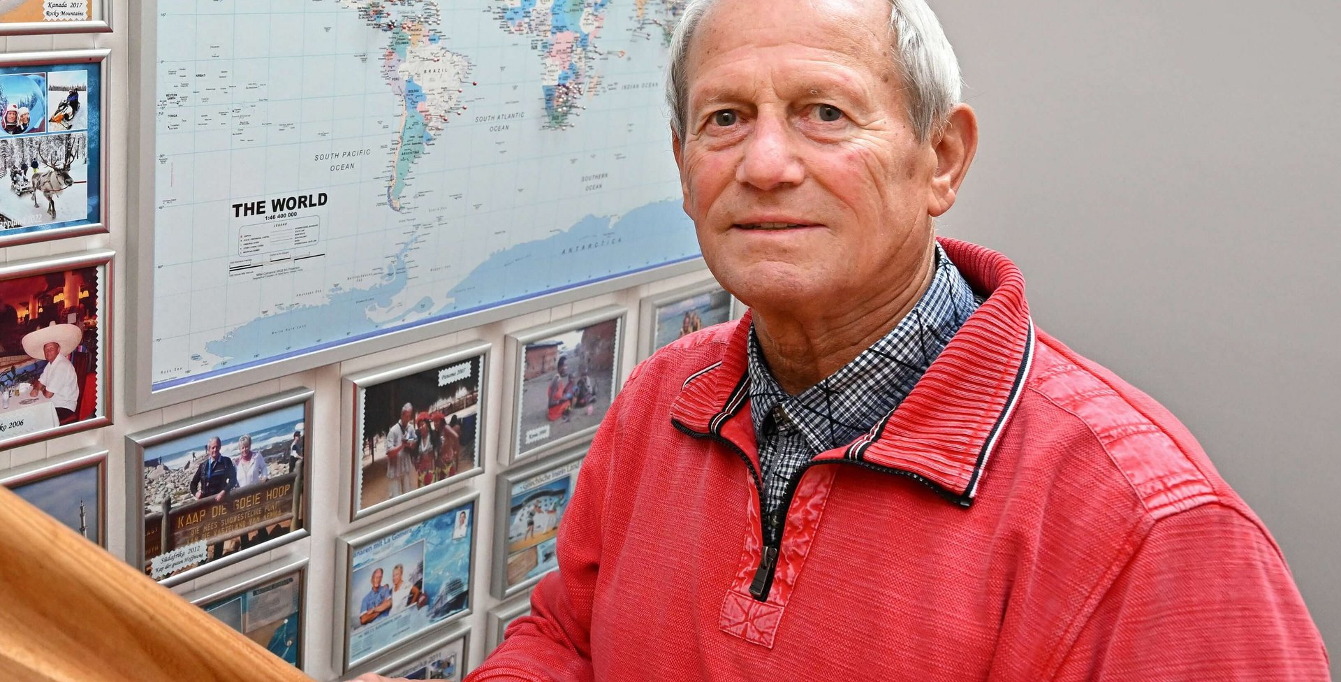 Lothar Schiller staat op een houten trap en houdt de leuning vast terwijl hij lacht. Op de achtergrond zijn talloze ingelijste foto's van zijn reizen te zien, die een wereldkaart omlijsten.