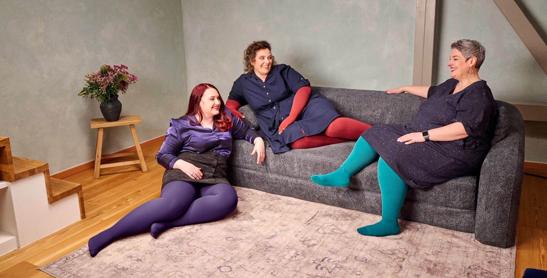 Drie vrouwen zitten ontspannen op een donkergrijze bank in een moderne woonkamer. Ze dragen kleurrijke panty's in paars, rood en turquoise bij donkere jurken of rokken en kletsen vrolijk met elkaar.