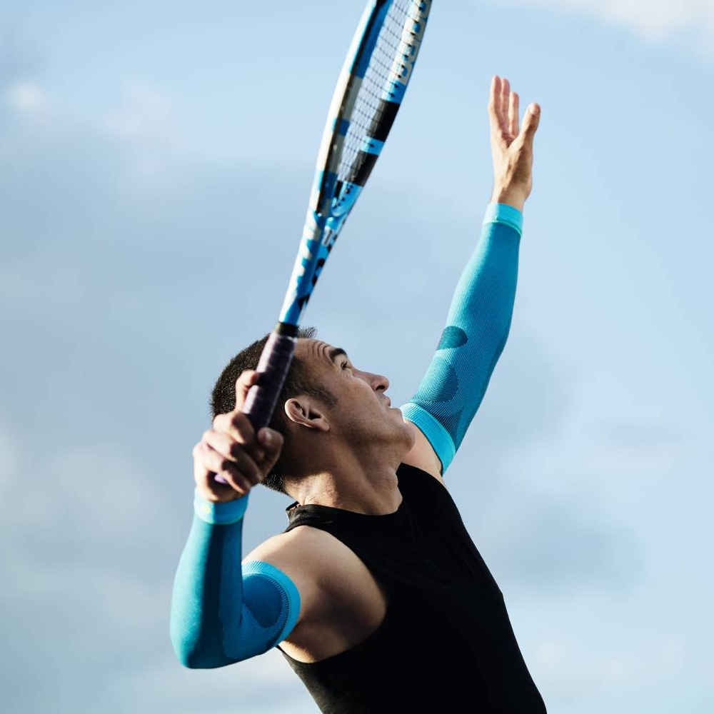 Een tennisser met blauwe compressiemouwen serveert. De lucht is blauw met enkele wolken.
