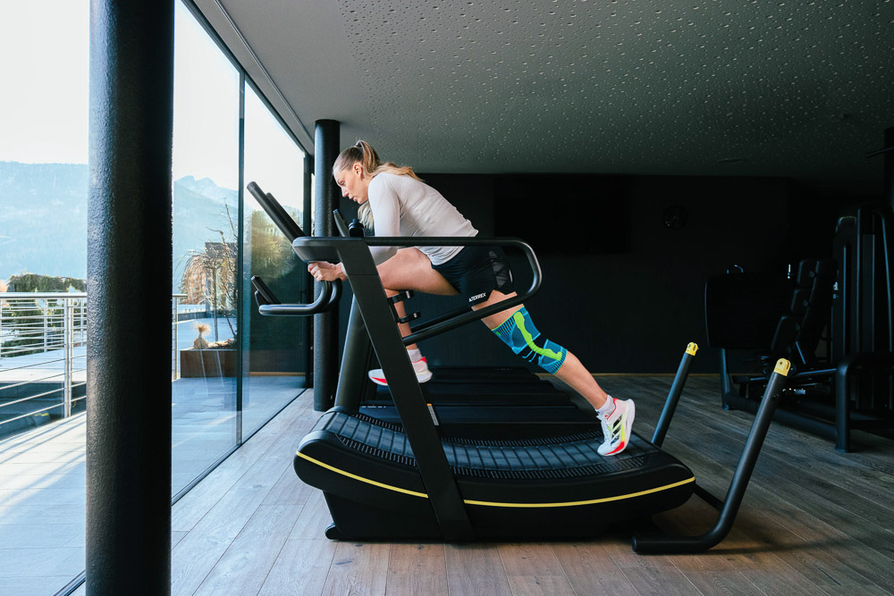 Sportieve vrouw voert een gecontroleerde uitvalspas uit met een dumbbell in haar hand. Ze traint in een moderne fitnessruimte met uitzicht op de bergen en draagt een kniebandage aan het voorste been voor extra stabiliteit tijdens de krachtoefening.