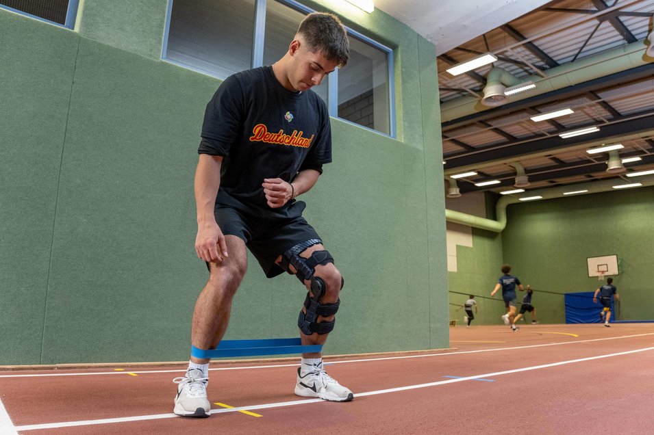 Simon Liedtke, professioneel honkballer, doet oefeningen in een gymzaal. Hij draagt een SecuTec Genu Flex knieorthese om zijn gescheurde achterste kruisband te stabiliseren. Op de achtergrond zie je andere spelers trainen.
