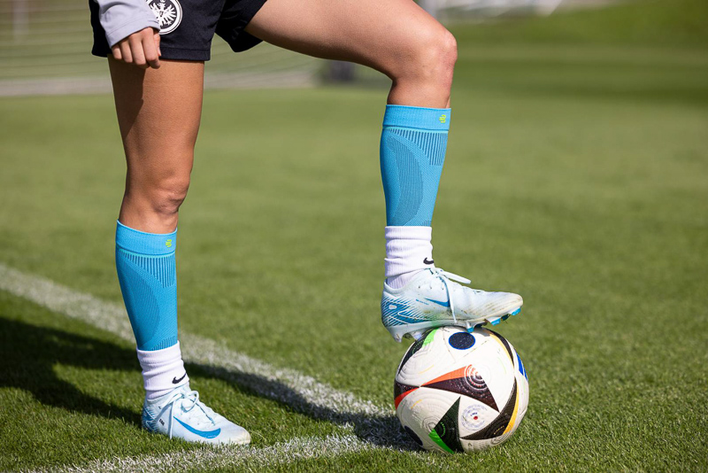 Close-up van een voetbalspeelster die Bauerfeind compressiekousen draagt, terwijl ze met één voet op een bal rust. Het groene veld en de scherpe details benadrukken de atletische prestatie en de hoogwaardige uitrusting.