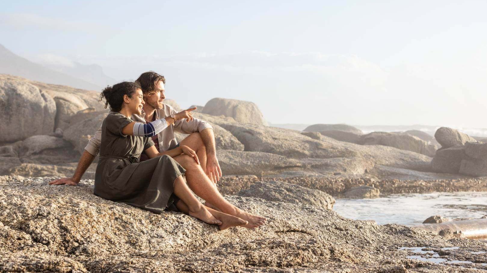 De afbeelding toont een stel dat op het strand zit. De vrouw draagt een EpiTrain elleboogbandage van Bauerfeind, die samen met regelmatige onderarmoefeningen kan helpen om elleboogpijn te voorkomen.