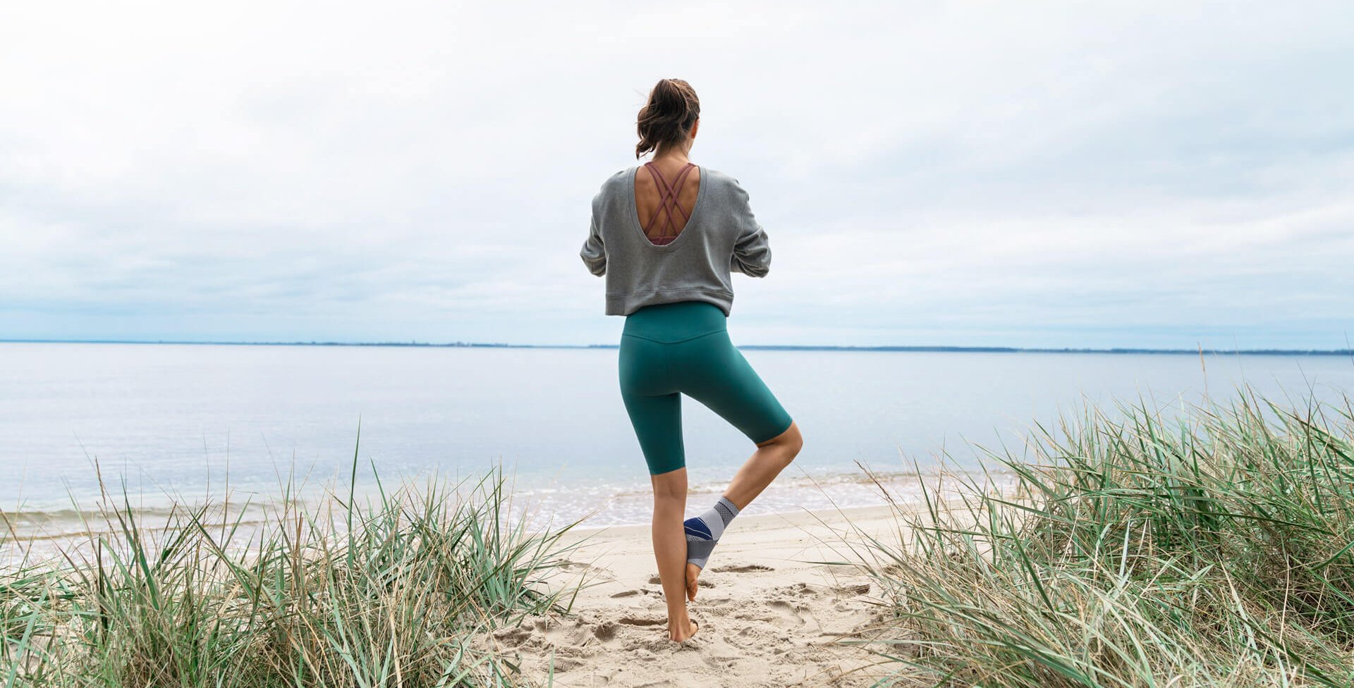 De foto toont een vrouw die op het strand een yogahouding aanneemt. Ze draagt de MalleoTrain enkelbandage van Bauerfeind om haar enkel te stabiliseren. Die komt onder meer van pas bij een enkeldistorsie.