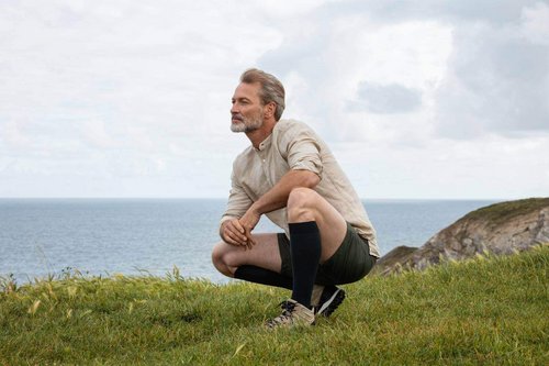 Een man hurkt op een weide aan zee. Hij draagt compressiekousen van Bauerfeind.