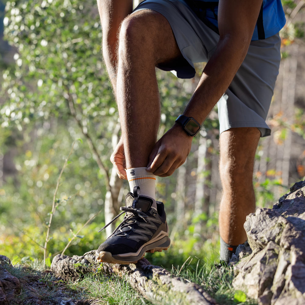 Een wandelaar staat op een smal bospad en trekt de Outdoor Compression Ankle Support om zijn enkel. Hij draagt stevige wandelschoenen, een korte wandelbroek en een rugzak.