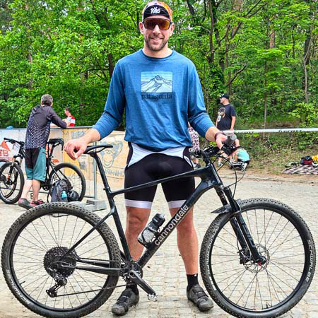 Een glimlachende man staat met een zwarte Cannondale-gravelbike op een bospad. Hij draagt een blauwe Patagonia trui met lange mouwen en een zwarte fietsbroek. Andere fietsers en groene bomen zijn op de achtergrond zichtbaar.