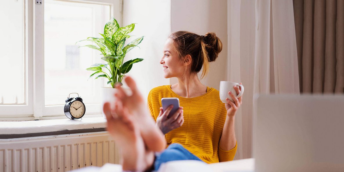 Op de foto zit een jonge vrouw op een stoel met haar voeten op de tafel voor haar. In haar linkerhand houdt ze een kopje, in haar rechterhand een smartphone. Regelmatig trainen van de voetspieren kan pijn voorkomen.