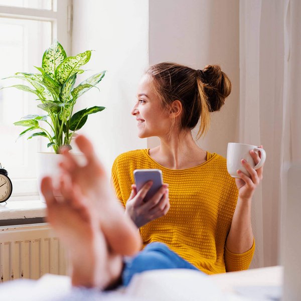 Op de foto zit een jonge vrouw op een stoel en heeft haar voeten op de tafel voor haar gelegd. In haar linkerhand houdt ze een kopje; in haar rechterhand een smartphone. Regelmatige training van de voetmusculatuur kan pijn voorkomen.